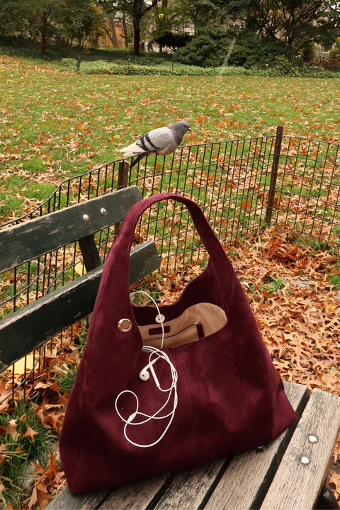 Maroon handbag with a shoe on top, placed on a wooden bench in a park with fallen leaves and a pigeon in the background.