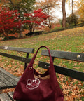 Maroon handbag with a white design on a wooden bench in a park with autumn leaves and trees.