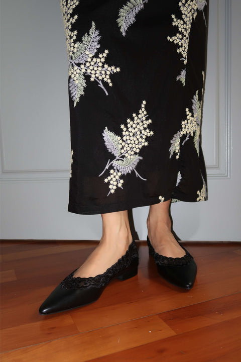 Person wearing a black floral embroidered skirt and black shoes on a wooden floor.
