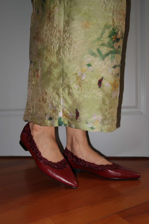 Person wearing a green floral skirt and red shoes on a wooden floor.